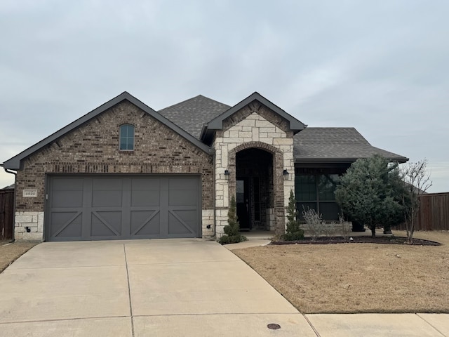 french country style house with stone siding, concrete driveway, brick siding, and roof with shingles