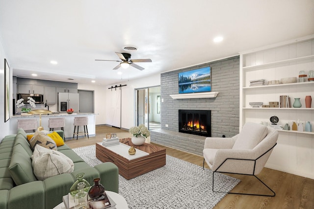 living room with a brick fireplace, a barn door, ceiling fan, and light hardwood / wood-style flooring