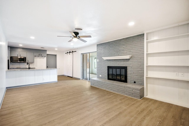 unfurnished living room with a brick fireplace, light hardwood / wood-style flooring, built in features, ceiling fan, and a barn door