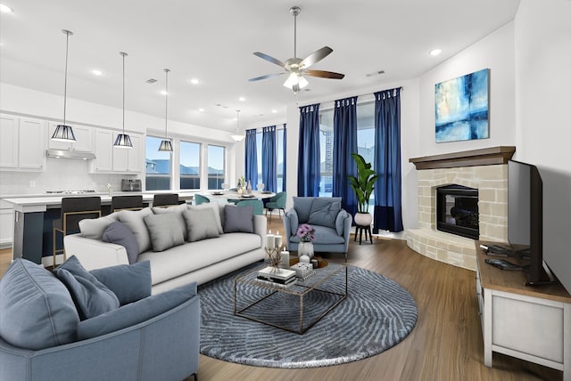 living room with a stone fireplace, dark wood-type flooring, and ceiling fan