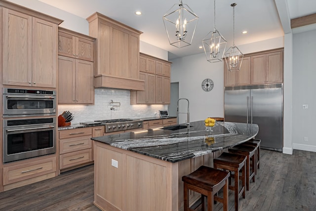 kitchen with appliances with stainless steel finishes, decorative light fixtures, an island with sink, sink, and dark stone countertops