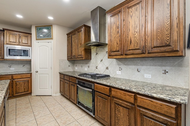 kitchen featuring wall chimney range hood, appliances with stainless steel finishes, light stone counters, tasteful backsplash, and light tile patterned flooring