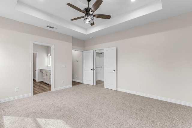 unfurnished bedroom with ceiling fan, connected bathroom, dark carpet, a walk in closet, and a raised ceiling