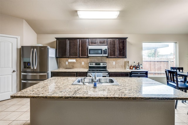 kitchen featuring light stone counters, dark brown cabinets, a center island with sink, light tile patterned floors, and appliances with stainless steel finishes