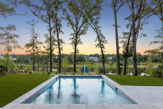 pool at dusk featuring a patio, a water view, and a lawn