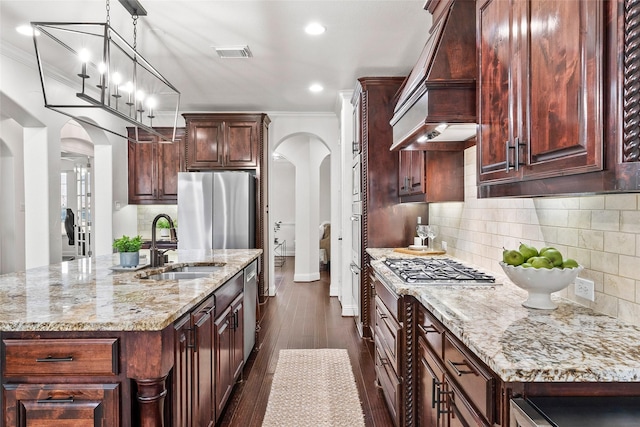 kitchen with premium range hood, appliances with stainless steel finishes, ornamental molding, a kitchen island with sink, and light stone counters