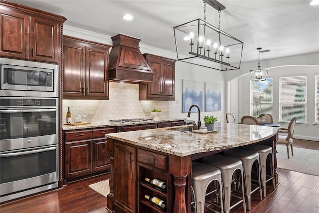 kitchen with appliances with stainless steel finishes, a kitchen island with sink, dark brown cabinets, custom range hood, and decorative light fixtures