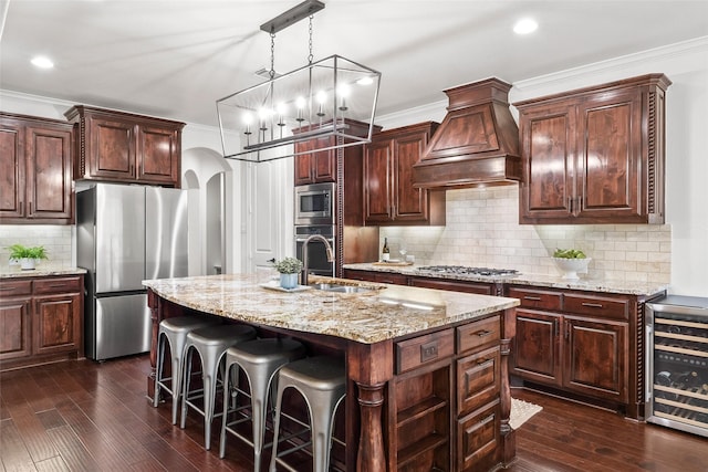 kitchen with a kitchen island with sink, ornamental molding, appliances with stainless steel finishes, and premium range hood