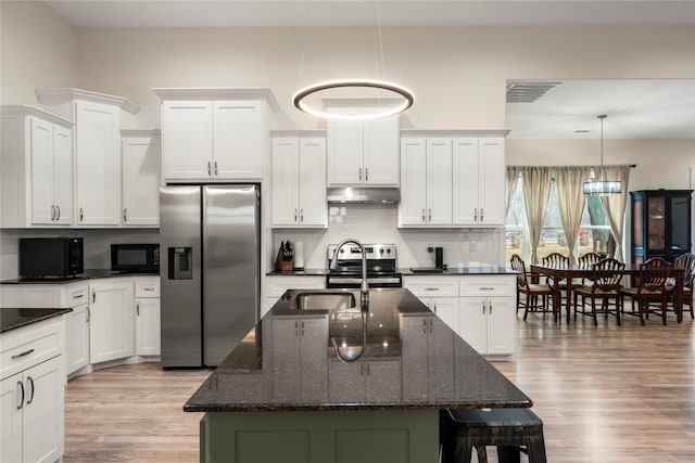 kitchen with appliances with stainless steel finishes, a kitchen island with sink, hanging light fixtures, dark stone counters, and light wood-type flooring