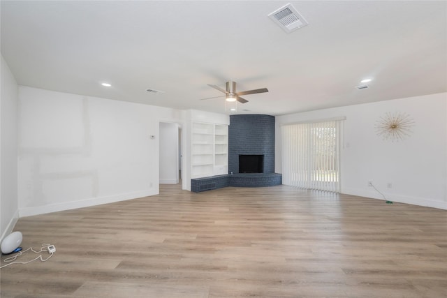 unfurnished living room with a brick fireplace, built in shelves, ceiling fan, and light wood-type flooring