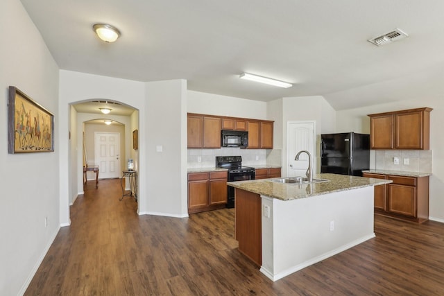 kitchen featuring sink, a kitchen island with sink, light stone counters, black appliances, and dark wood-type flooring