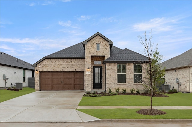 french country home with a shingled roof, central AC unit, concrete driveway, a front lawn, and brick siding