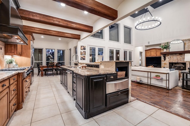 kitchen with an island with sink, beam ceiling, light stone countertops, sink, and island exhaust hood