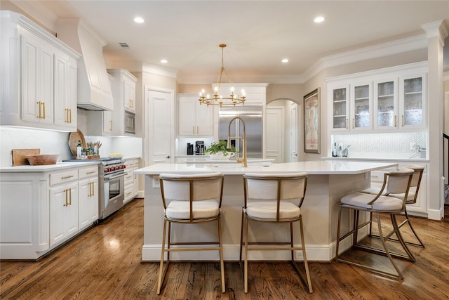 kitchen with pendant lighting, an island with sink, white cabinets, built in appliances, and dark wood-type flooring