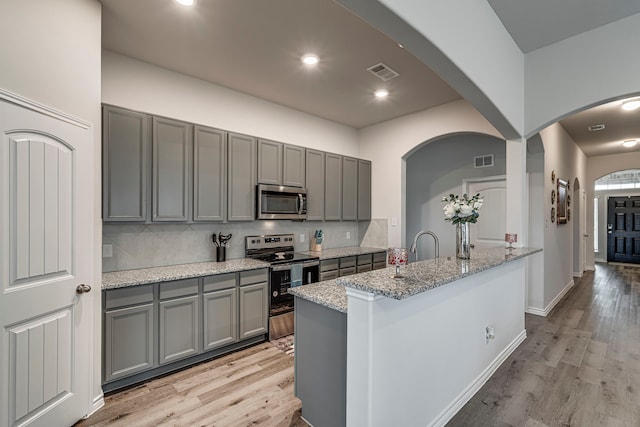 kitchen featuring stainless steel appliances, a kitchen island with sink, gray cabinetry, and light stone counters