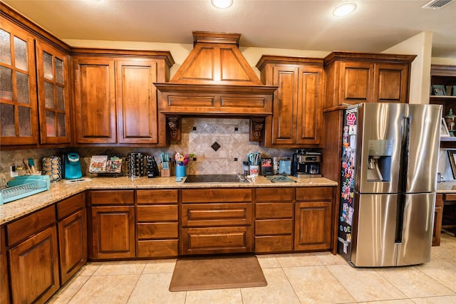 kitchen featuring stainless steel refrigerator with ice dispenser, light stone counters, black electric stovetop, custom range hood, and decorative backsplash