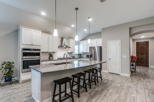 kitchen with sink, a kitchen island with sink, stainless steel appliances, white cabinets, and wall chimney exhaust hood