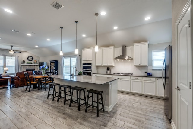 kitchen featuring white cabinetry, hanging light fixtures, a kitchen island with sink, stainless steel appliances, and wall chimney exhaust hood