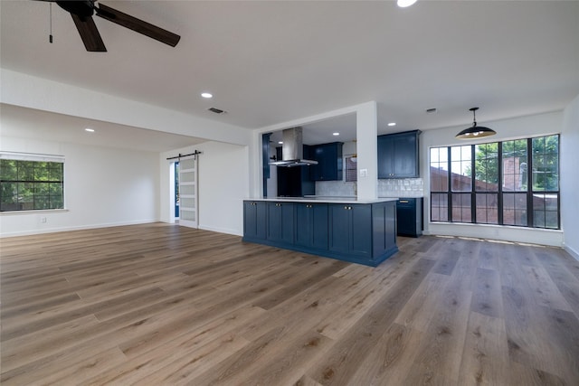 kitchen featuring blue cabinetry, decorative backsplash, a barn door, open floor plan, and wall chimney exhaust hood