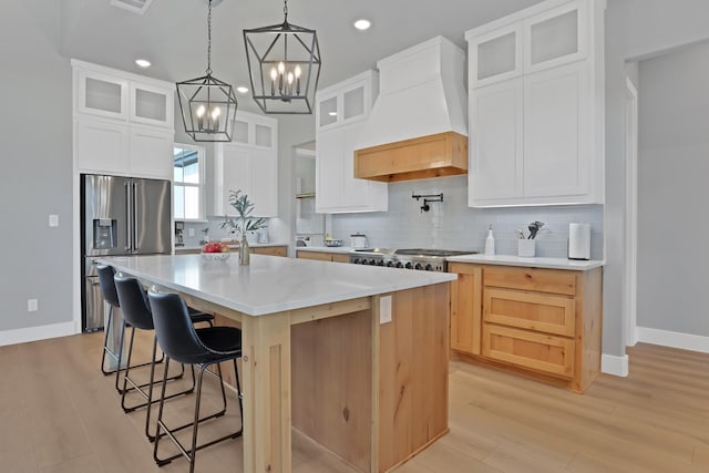 kitchen with high end fridge, custom range hood, an island with sink, white cabinets, and decorative backsplash