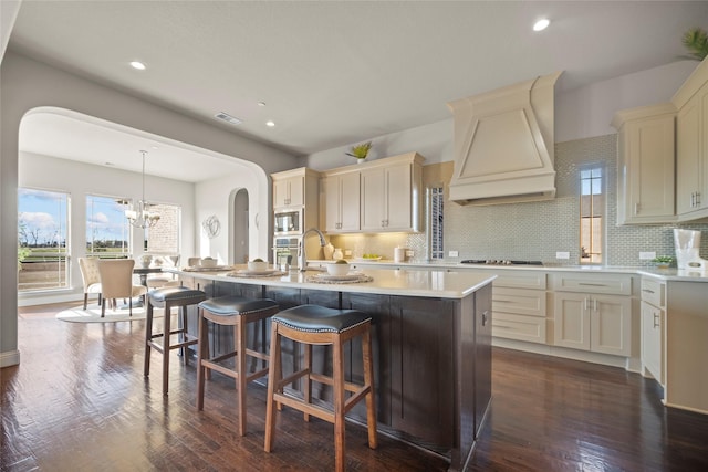 kitchen featuring dark wood-type flooring, custom exhaust hood, decorative light fixtures, stainless steel appliances, and a kitchen island with sink