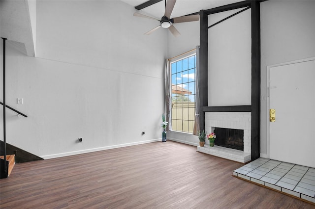 unfurnished living room with wood finished floors, a towering ceiling, baseboards, a ceiling fan, and a brick fireplace