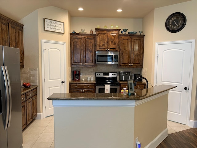 kitchen featuring light tile patterned floors, stainless steel appliances, dark stone counters, and a center island with sink