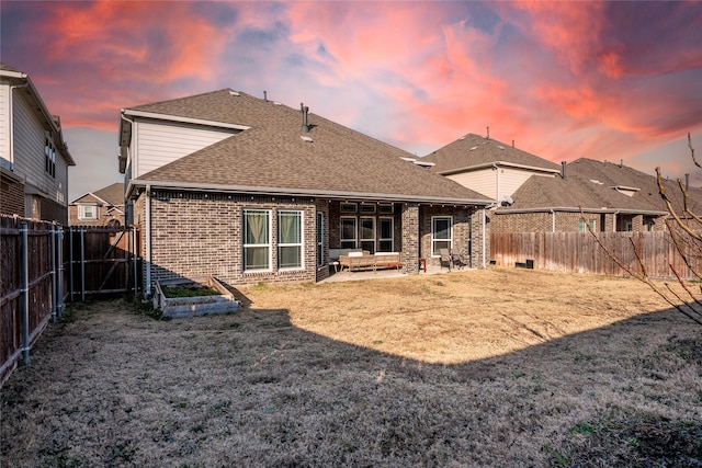 back of house at dusk with a patio area, brick siding, a fenced backyard, and roof with shingles