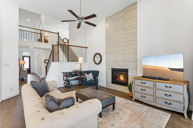 living room with a tiled fireplace, a towering ceiling, dark wood-type flooring, and ceiling fan