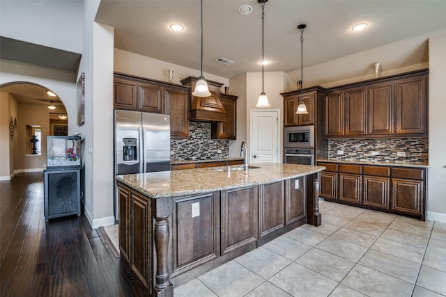 kitchen featuring decorative light fixtures, dark brown cabinets, stainless steel appliances, light stone countertops, and a kitchen island with sink