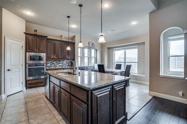 kitchen with an island with sink, sink, hanging light fixtures, stainless steel appliances, and light stone countertops