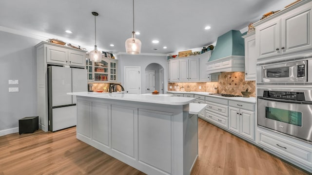 kitchen featuring decorative light fixtures, an island with sink, backsplash, stainless steel appliances, and custom range hood