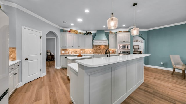 kitchen featuring crown molding, appliances with stainless steel finishes, an island with sink, custom range hood, and pendant lighting