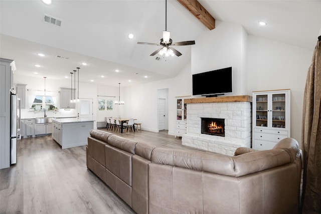 living room featuring sink, beam ceiling, a fireplace, ceiling fan with notable chandelier, and light wood-type flooring