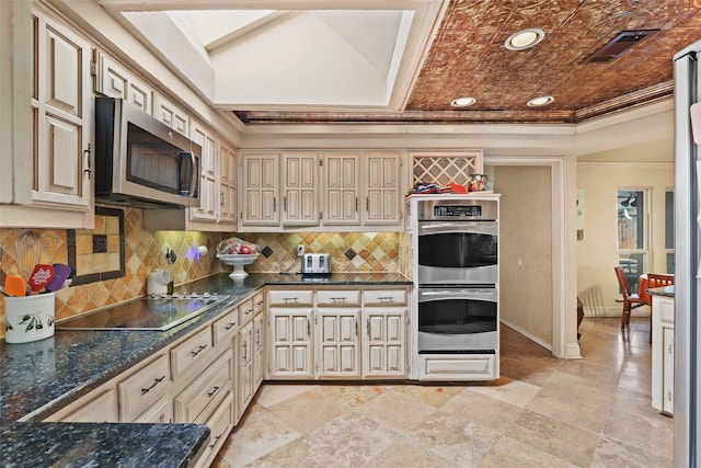 kitchen featuring crown molding, dark stone countertops, appliances with stainless steel finishes, cream cabinetry, and backsplash