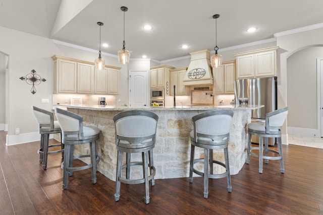 kitchen with dark wood finished floors, cream cabinets, appliances with stainless steel finishes, light stone countertops, and premium range hood