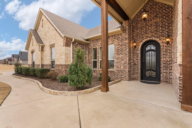 entrance to property featuring stone siding, roof with shingles, and brick siding