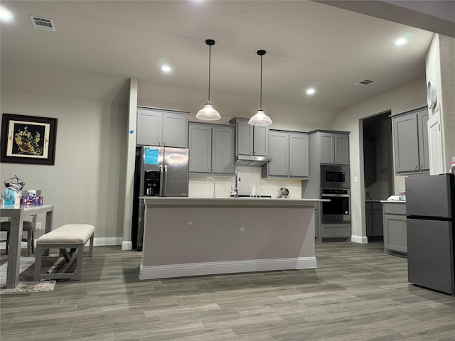 kitchen featuring tasteful backsplash, gray cabinetry, hanging light fixtures, stainless steel appliances, and a center island with sink