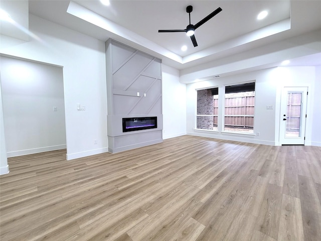 unfurnished living room featuring a large fireplace, a raised ceiling, ceiling fan, and light wood-type flooring