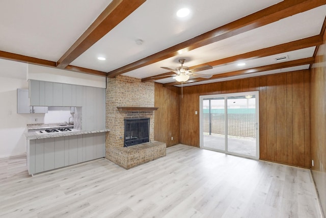 unfurnished living room with wooden walls, beamed ceiling, ceiling fan, a brick fireplace, and light wood-type flooring