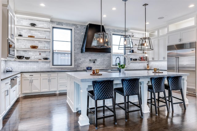 kitchen featuring a kitchen island with sink, hanging light fixtures, white cabinetry, a kitchen breakfast bar, and stainless steel built in refrigerator