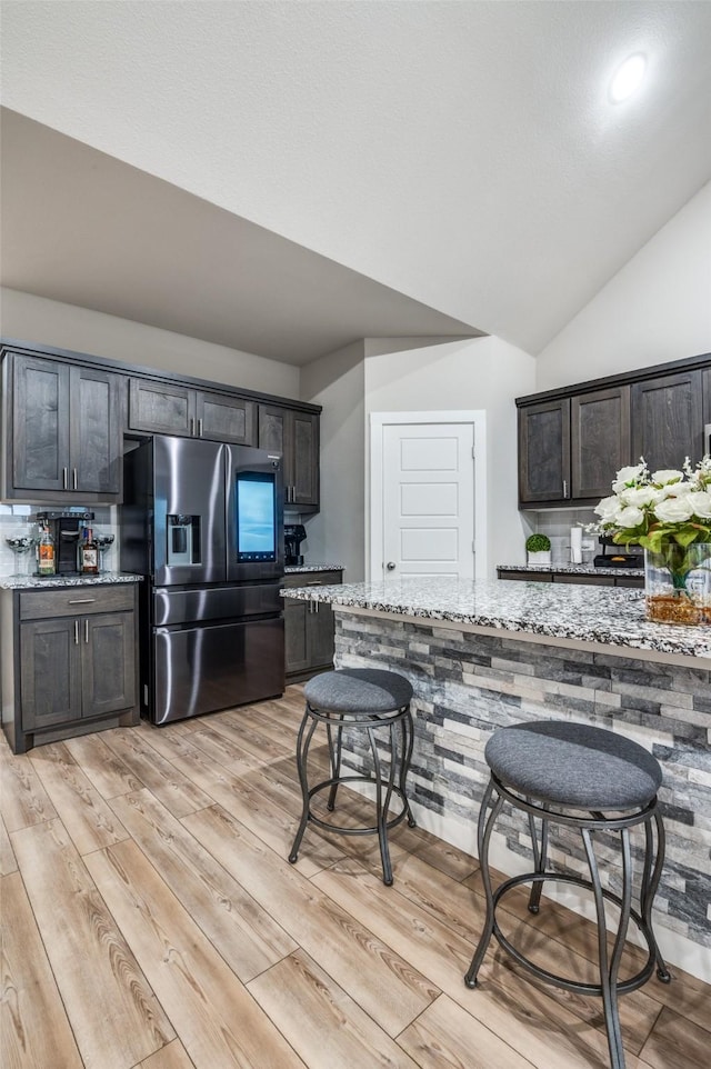 kitchen featuring stainless steel refrigerator with ice dispenser, lofted ceiling, dark brown cabinets, light wood-type flooring, and light stone countertops