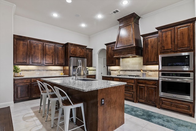 kitchen with premium range hood, sink, appliances with stainless steel finishes, light stone countertops, and a kitchen island with sink