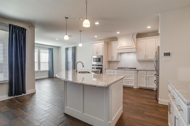 kitchen featuring premium range hood, appliances with stainless steel finishes, decorative light fixtures, an island with sink, and sink