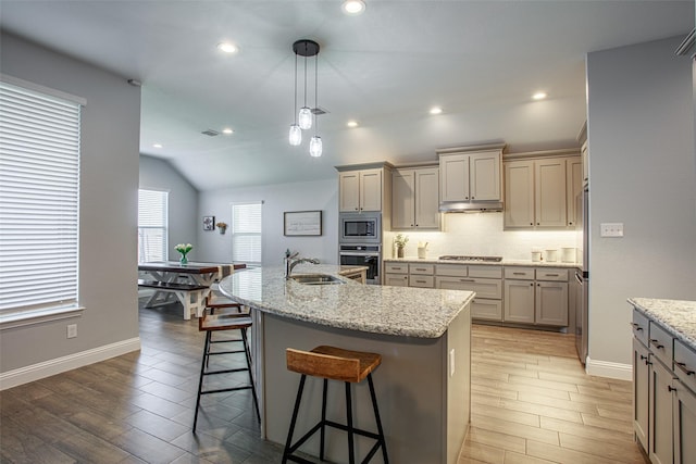 kitchen with backsplash, stainless steel appliances, light stone counters, an island with sink, and decorative light fixtures