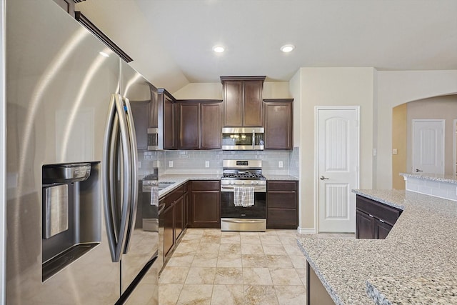 kitchen with dark brown cabinetry, light stone countertops, vaulted ceiling, appliances with stainless steel finishes, and decorative backsplash