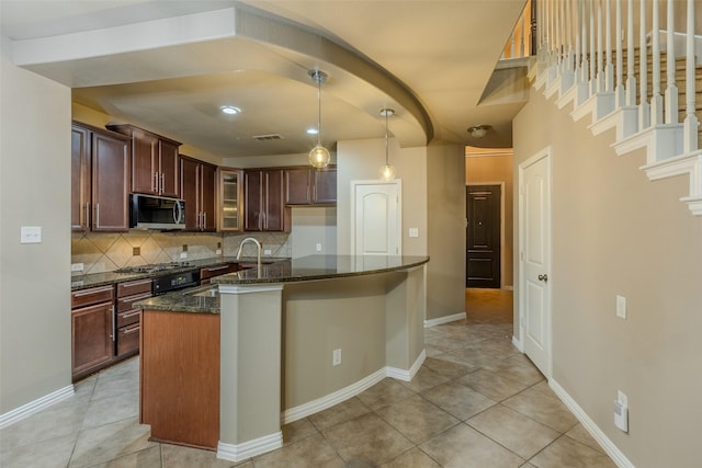 kitchen with appliances with stainless steel finishes, a kitchen island with sink, backsplash, hanging light fixtures, and dark stone counters