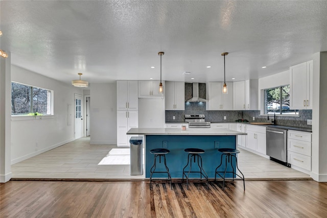 kitchen featuring a kitchen island, white cabinetry, hanging light fixtures, stainless steel appliances, and wall chimney range hood