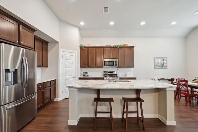 kitchen with dark hardwood / wood-style flooring, stainless steel appliances, light stone countertops, a kitchen island with sink, and decorative backsplash