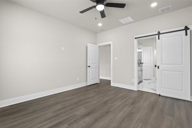 unfurnished bedroom featuring dark hardwood / wood-style floors, ensuite bath, a barn door, and ceiling fan